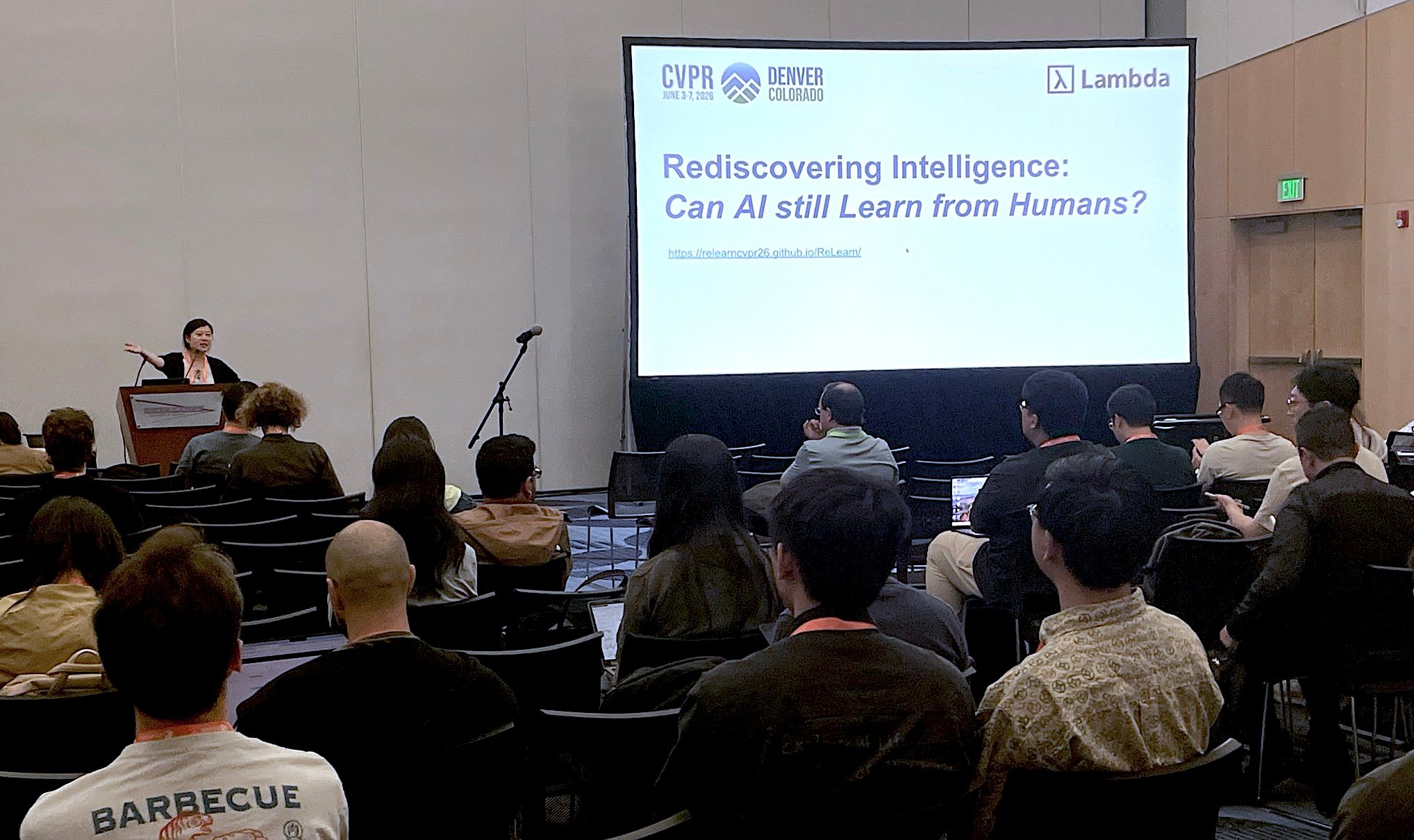 A speaker at a podium opens the ReLearn workshop at CVPR 2026 in Denver, gesturing toward a large screen that reads "Rediscovering Intelligence: Can AI still Learn from Humans?" with CVPR and Lambda logos. An audience fills rows of chairs in the foreground.